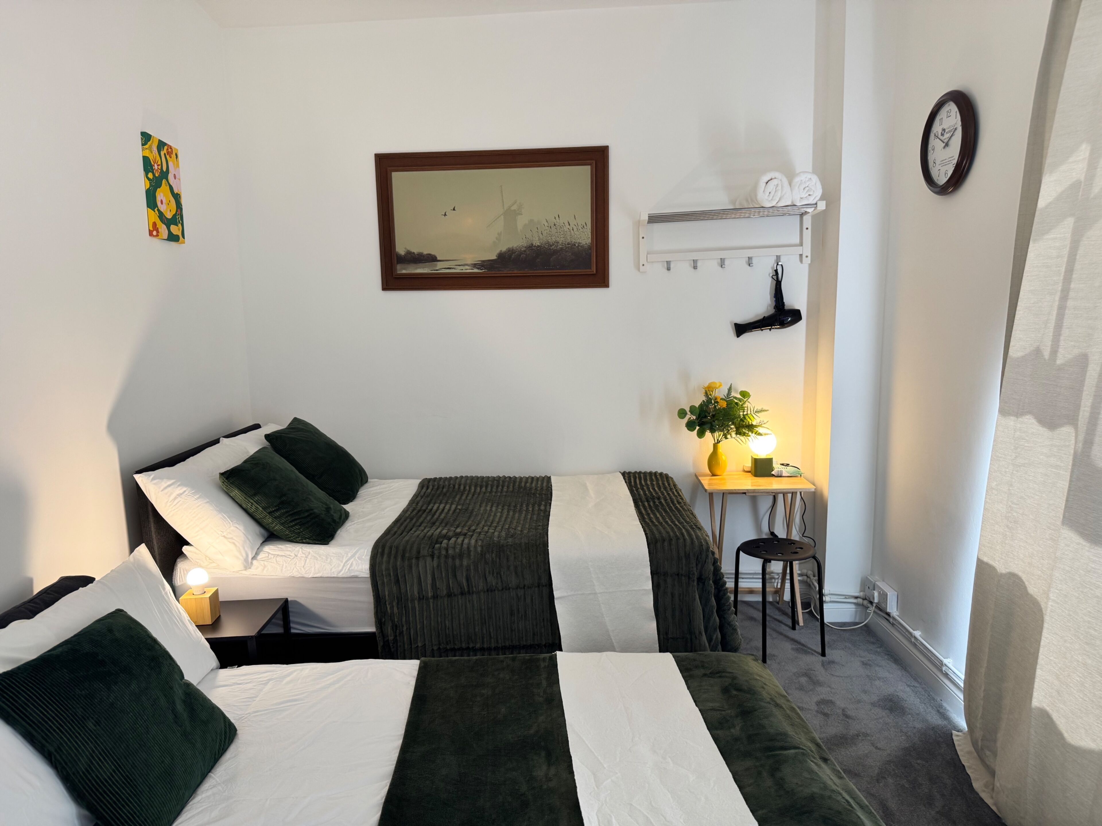 A hotel room with two beds, a painting, a clock, a small table with a lamp, and a hairdryer on the wall.