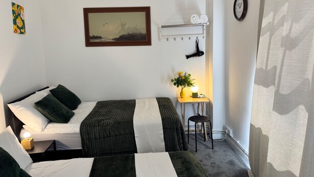 A room with two beds, a desk with a lamp, a painting, a clock, and a shoe rack. - Eurostar Private Flat Sleeps up to 4 London (London)