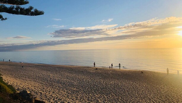 Een strand met mensen die langs de kust lopen, een heldere lucht en de zon die in het water ondergaat of opkomt.