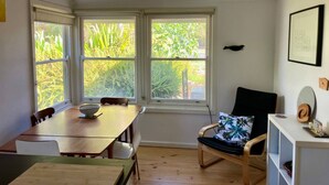 Een eethoek met een houten tafel en stoelen, grote ramen met uitzicht op groen, en een witte muur met een lijst en een klein plankje.