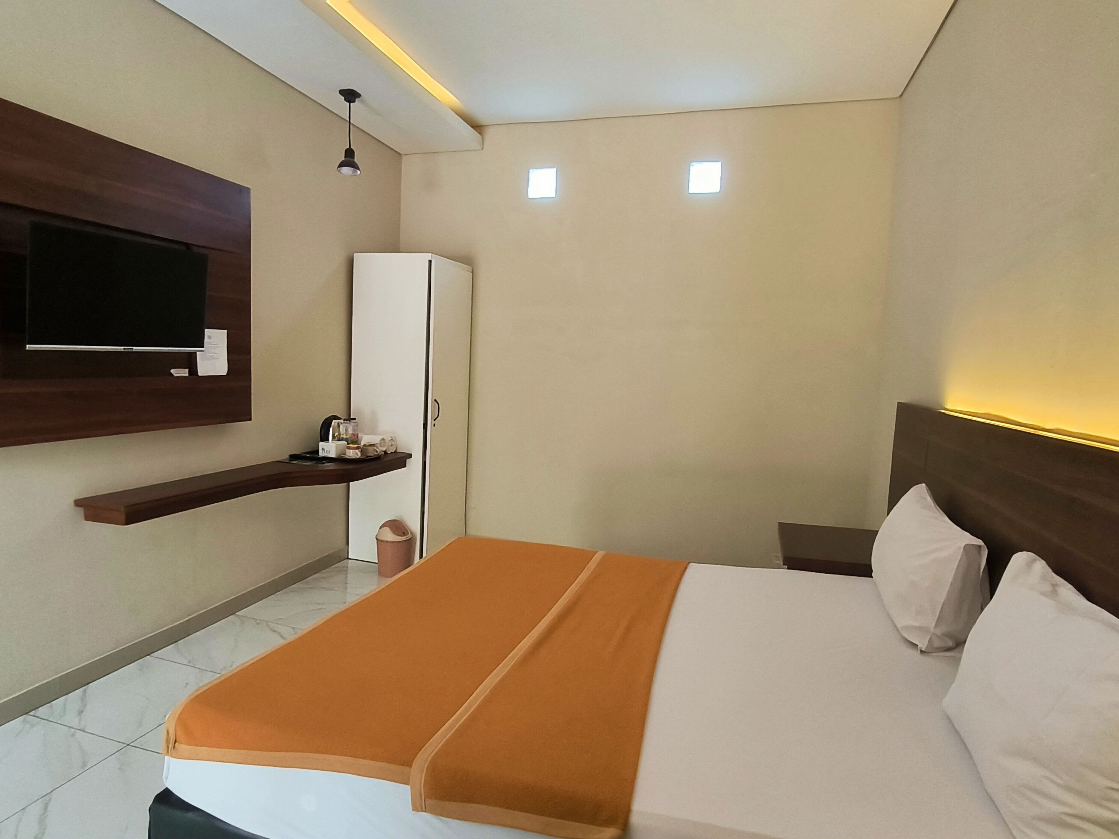 A hotel room with a bed, a TV mounted on the wall, a small cabinet, and a shelf with toiletries.