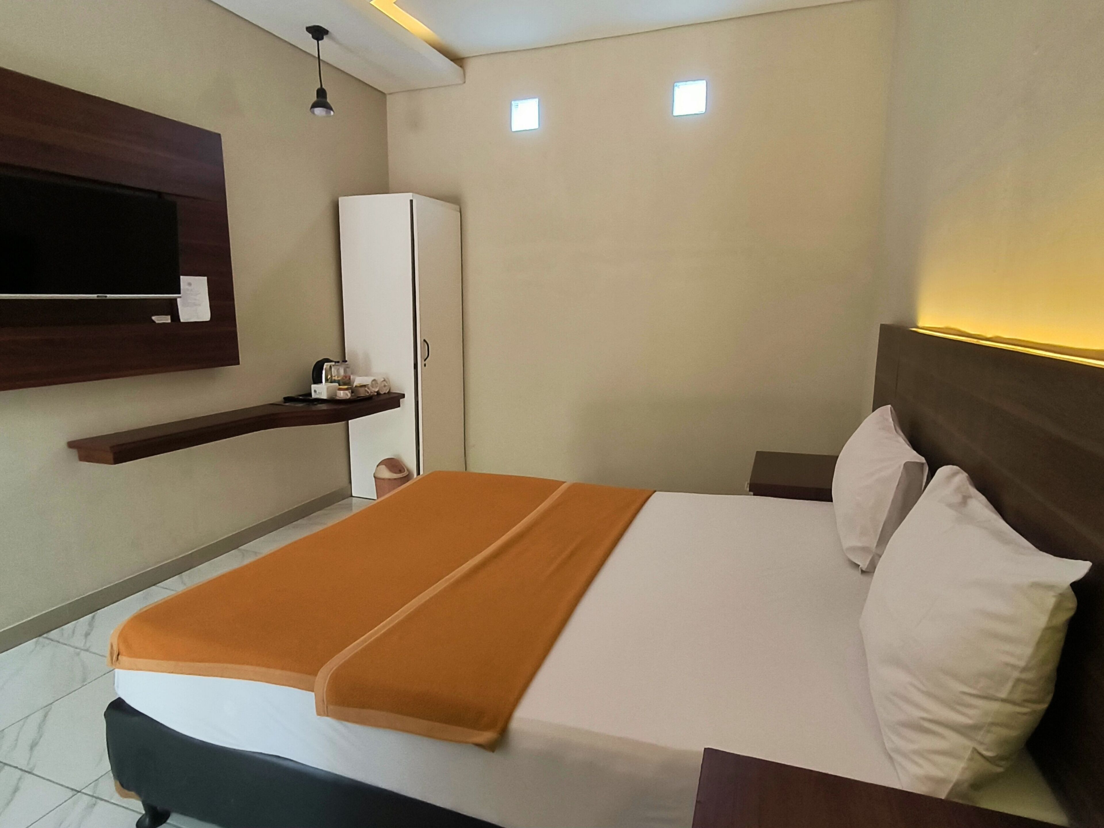 A hotel room with a bed, a TV mounted on the wall, a wardrobe, and a small shelf with toiletries.