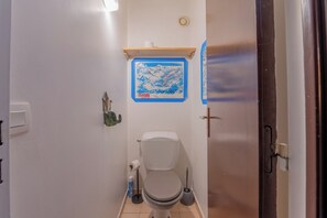 Un baño pequeño y funcional con inodoro, lavamanos y zona de ducha. Hay un perchero en la pared y un estante pequeño sobre el inodoro.