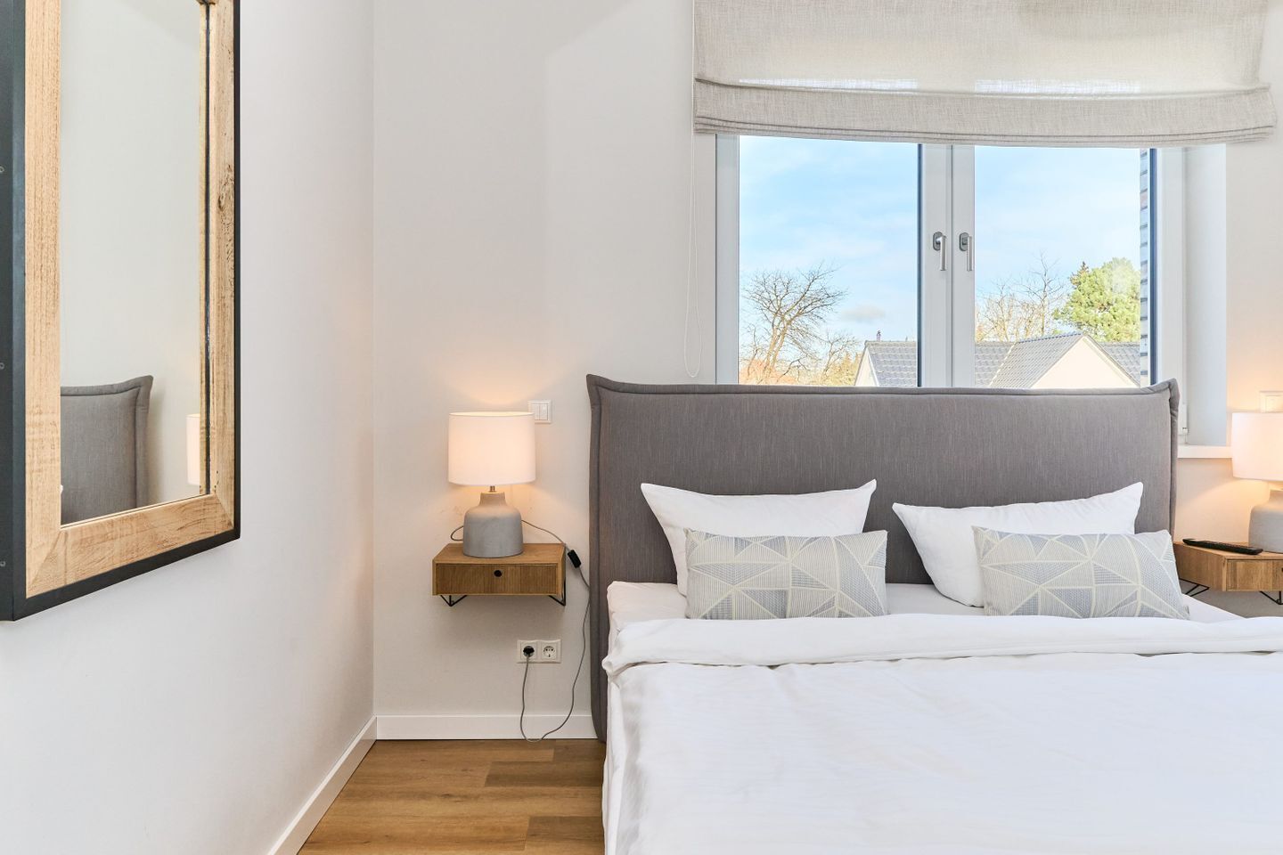 Een slaapkamer met een bed, twee nachtkastjes met lampen, een groot raam met uitzicht op bomen en een spiegel aan de muur.