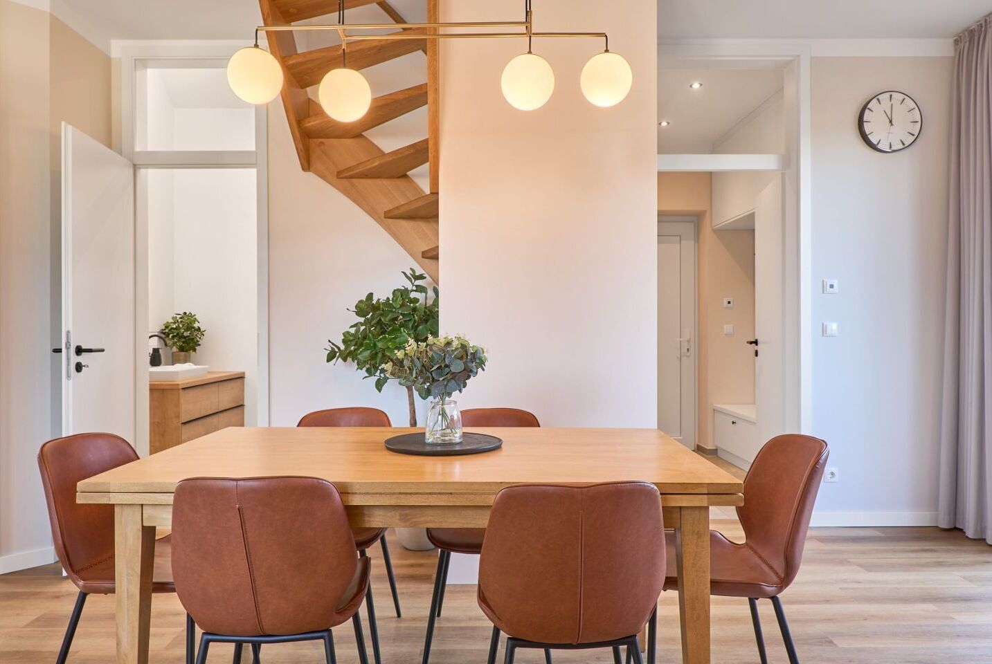 Zona pranzo moderna con tavolo in legno, sei sedie marroni, un vaso con fiori e una scala a chiocciola con luci a sospensione.