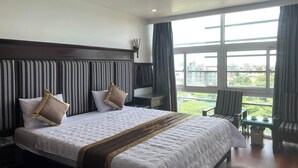 Habitación de hotel con una cama grande, vistas a la ciudad a través de amplios ventanales y una pequeña mesa con una silla.