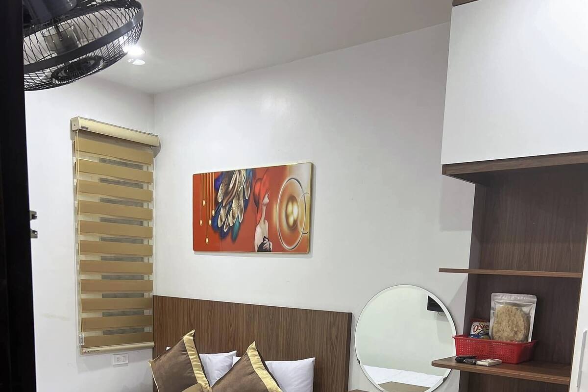 Ein kleines Hotelzimmer mit Bett, Schreibtisch, kleinem Kühlschrank, Spiegel und einem Wandbild.