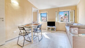 Ett kompakt bostadsutrymme med en matplats, en platt-TV, en soffa och utsikt över byggnader genom fönstren.