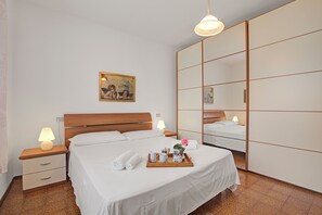 Ein ordentlich bezogenes Bett mit weißen Leinen, ein Nachttisch mit Lampe, ein Tablett mit Handtüchern und einer Blumenvase sowie eine große Spiegelschränke.