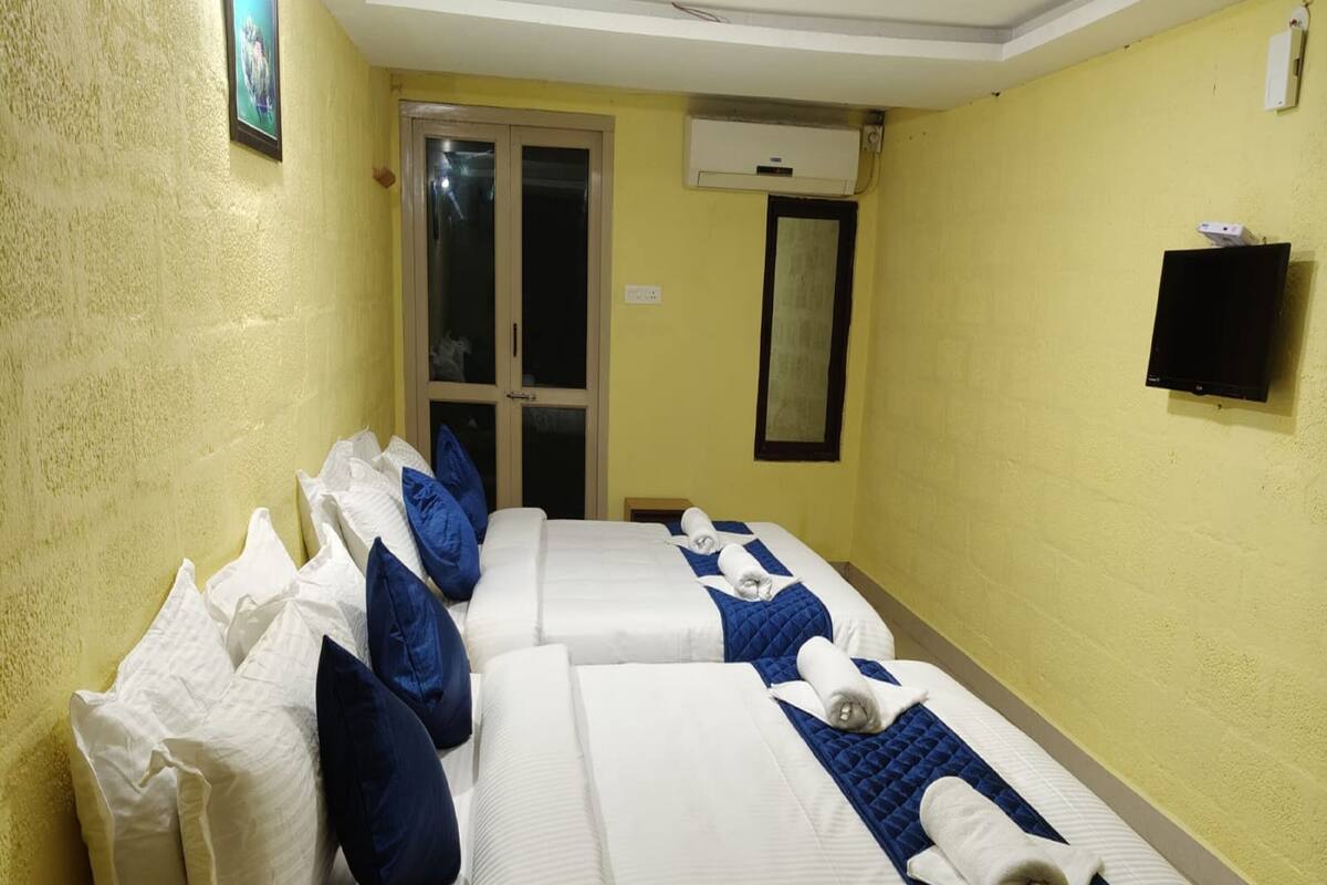 A hotel room with two beds, each with white bedding and blue accents, a small TV mounted on the wall, and an air conditioning unit above the door.