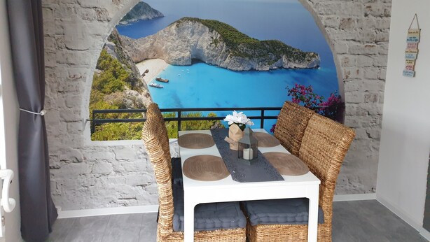 Comedor con vistas a un paisaje costero a través de un arco de piedra, muebles de mimbre y una mesa con vela y adornos.