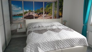 Un dormitorio con cama, mesita de noche, lámpara, ventana con vista a una playa y un mural tropical en la pared.