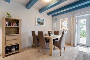 A dining area with a wooden table, six chairs, a bookshelf, a clock, and a window with curtains. - Apartment "Vogelkoje" with Private Terrace, Shared Garden and Wi-Fi (Pellworm)