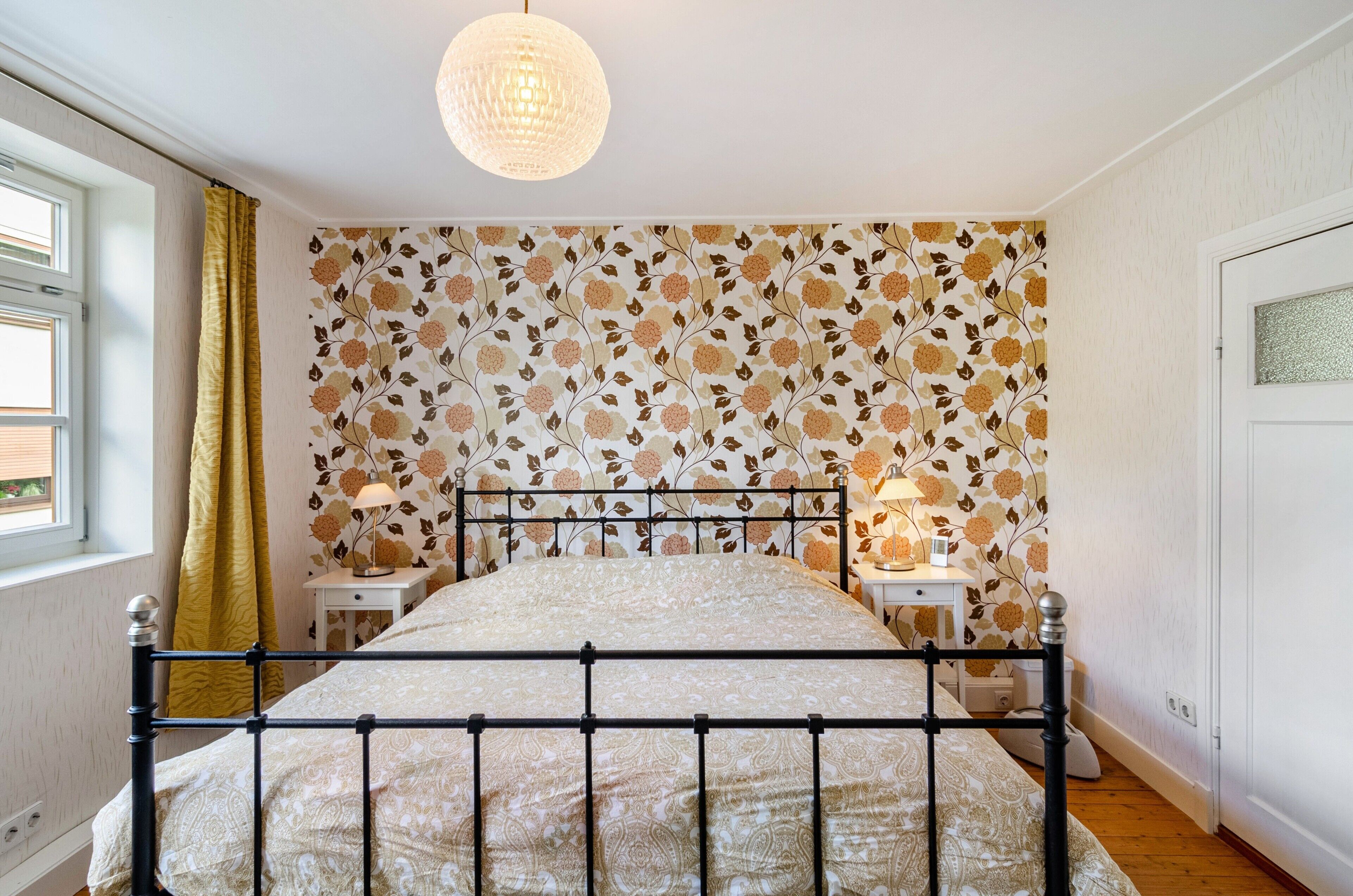A bedroom with a bed, a nightstand, a lamp, a window with curtains, and a wall with floral wallpaper.