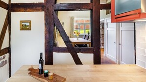 Une pièce avec des poutres apparentes, une table en bois sur laquelle se trouvent une bouteille et un verre, un téléviseur fixé au mur, et une vue sur un coin repas à travers une fenêtre.