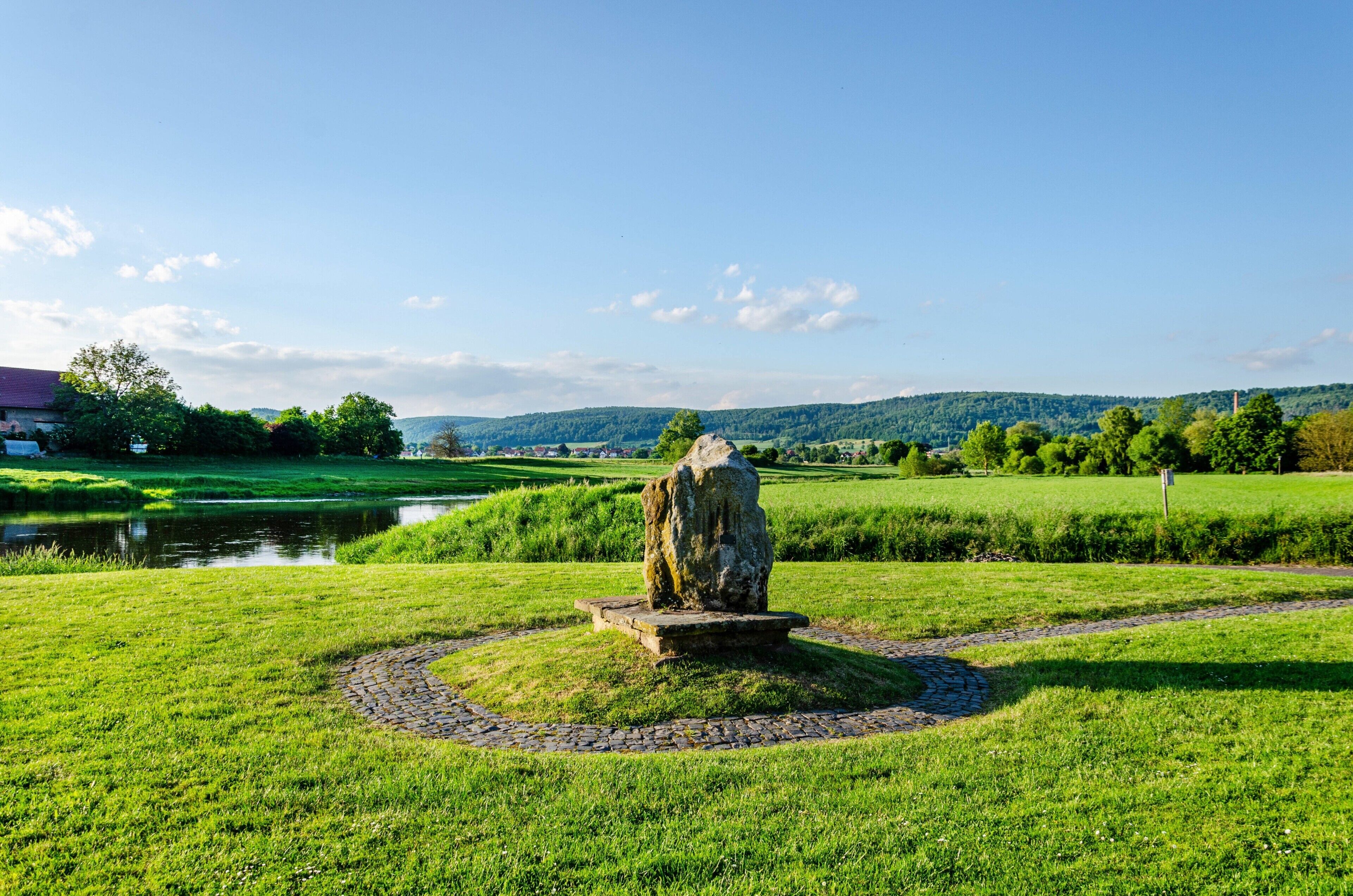 A stone monument in a grassy field with a river and hills in the background.