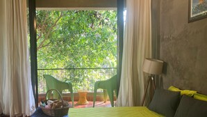 Quarto com cama, janela com cortinas, uma cadeira, abajur e vista para o verde lá fora.
