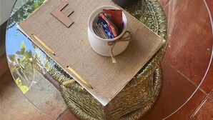 Uma mesa de vidro redonda com um descanso trançado, uma tigela branca pequena com cabo e uma planta em vaso com flores roxas.