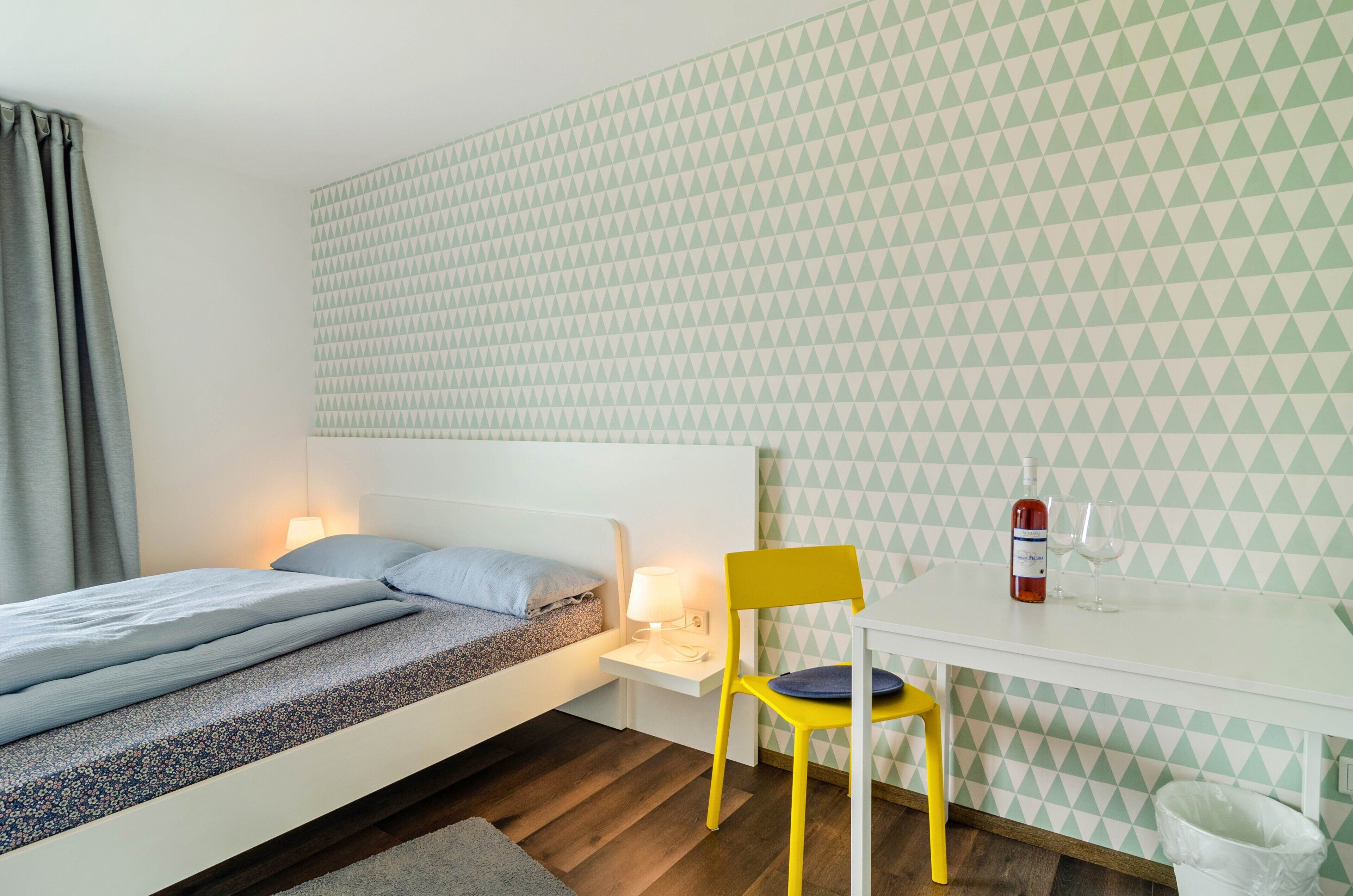 Une chambre avec un lit, un bureau avec une chaise, une lampe et une bouteille d’eau.