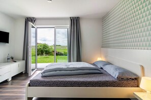 A modern bedroom with a bed, bedside table, and a view of a green landscape through a large window. - Room 'Doppelzimmer' with Private Terrace, Shared Garden and Wi-Fi (Leibertingen)