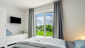 A hotel room with a bed, a TV mounted on the wall, a desk with a water bottle and plant, and a window with curtains. - Room 'Doppelzimmer' with Private Terrace, Shared Garden and Wi-Fi (Leibertingen)