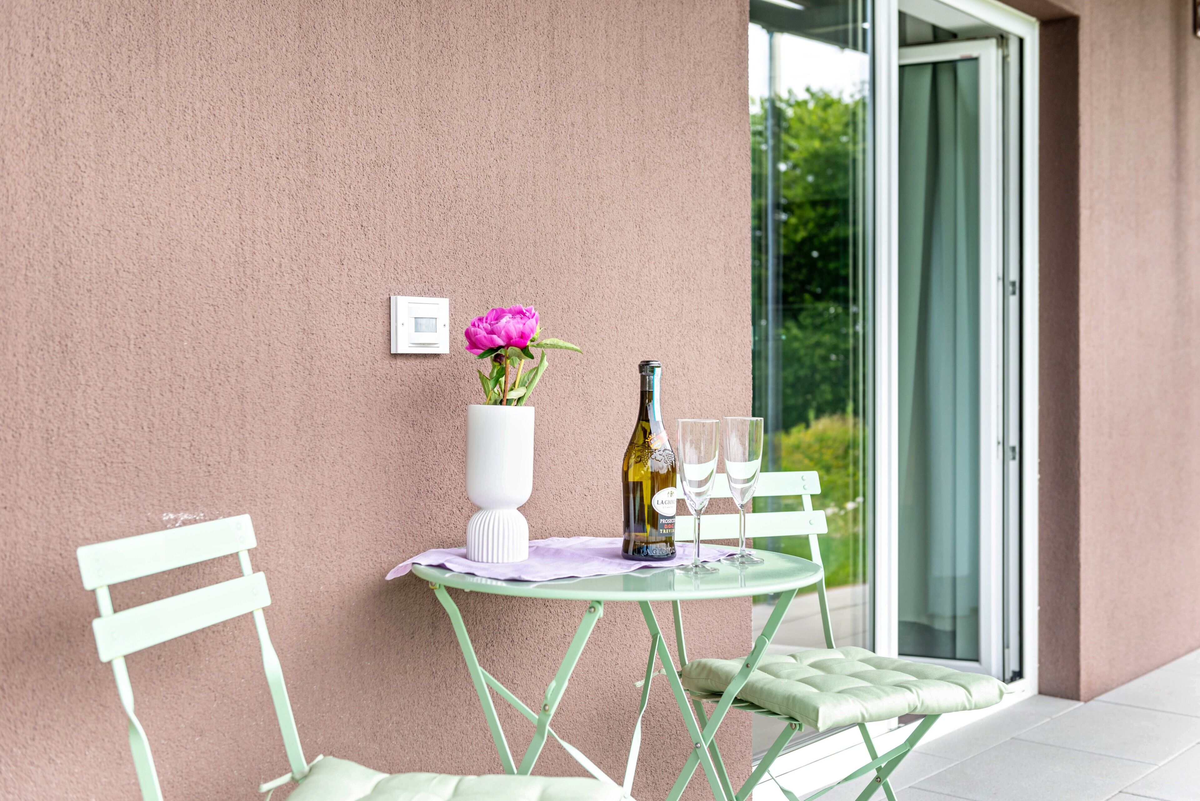 Une table sur laquelle se trouvent un vase de fleurs, une bouteille et deux verres, le tout posé sur une table en verre, avec une chaise blanche à côté.