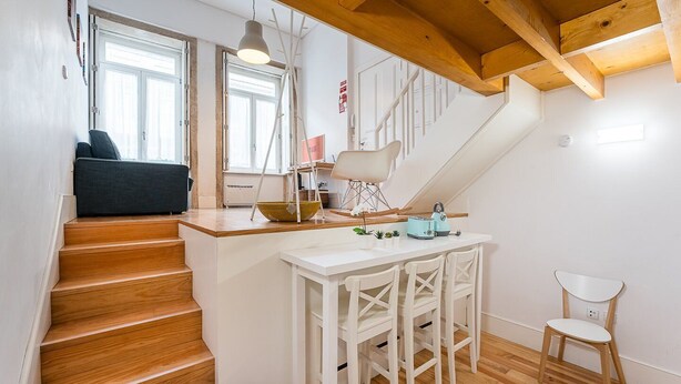 A modern kitchen with a wooden staircase, white bar stools, and a small dining table.