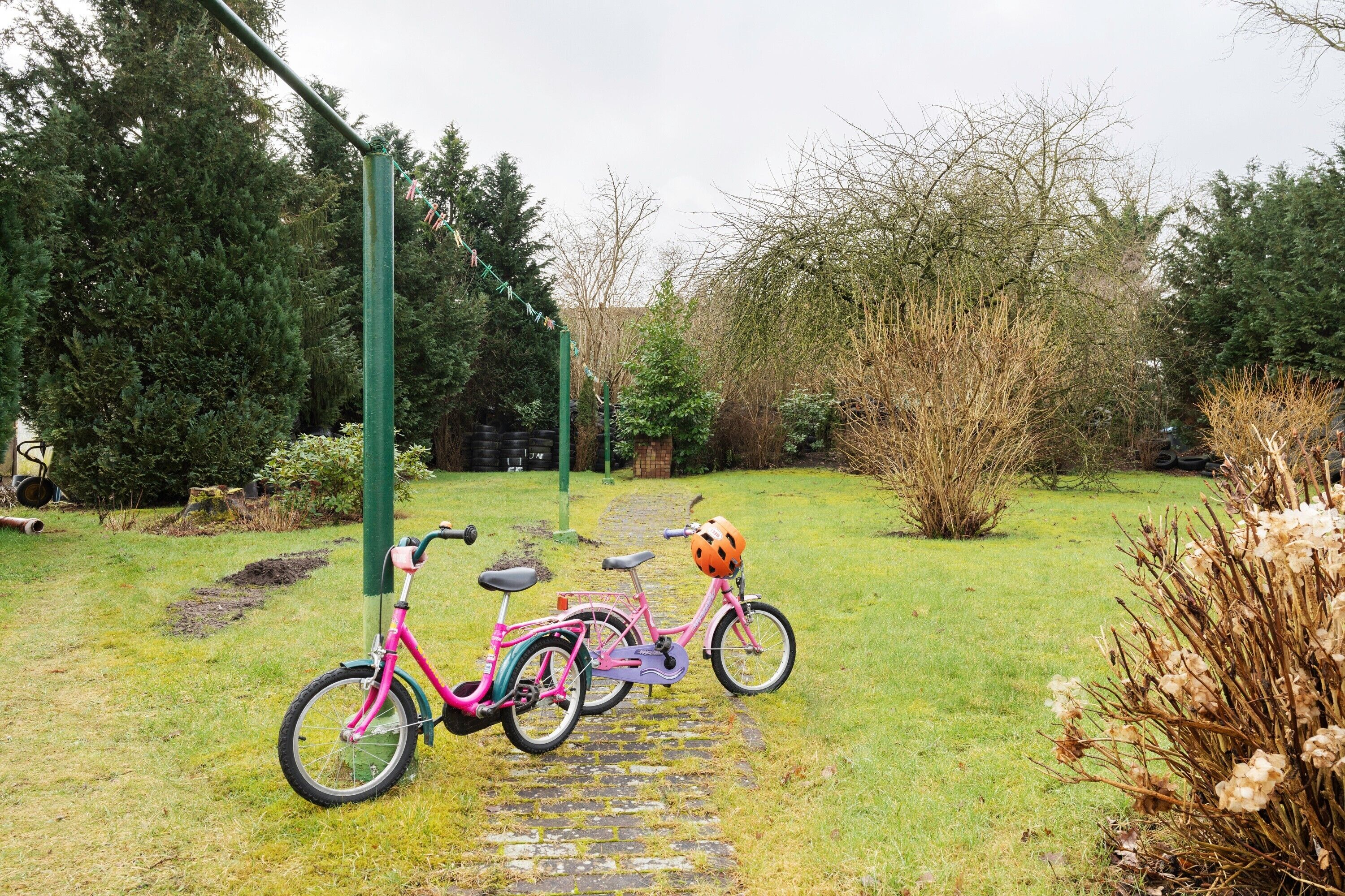 Ein pinkfarbenes Fahrrad mit Helm und Korb, abgestellt auf einem steinbelagten Weg in einem Garten mit Bäumen und Sträuchern.