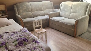 Una habitació amb un llit, dues butaques, un tamboret de fusta i un aparell de ràdio antic.