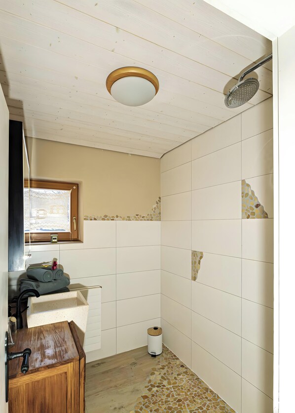Un bagno con un mobile bagno in legno, una doccia con soffione a mano, un water e una finestra con persiane.
