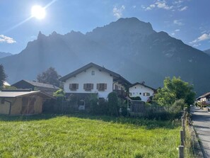 Un paysage de montagne avec un bâtiment blanc doté de volets en bois, entouré de verdure et sous un ciel dégagé.