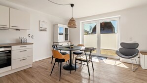 A modern kitchen with white cabinets, a dining table with chairs, and a large window with blinds. - Apartment 'Ferienhaus Waterkant Mit Hund' with Private Terrace, Private Garden and Wi-Fi (Klanxbüll)