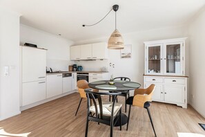 Moderni keittiö, jossa on valkoiset kaapit, ruokapöytä tuoleineen, riippuvalaisin ja lasiovilla varustettu kaappi.