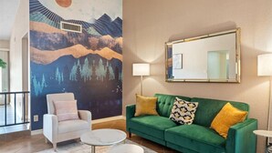 A living room with a green sofa, a white armchair, and a round coffee table. There is a large mural on the wall depicting a mountainous landscape. - Desert Paradise Pool, Mini Golf, Fire Pit, Games, Relax and unwind (Phoenix)
