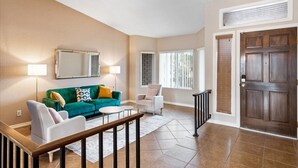 A living room with a green sofa, a white armchair, and a coffee table. There is a wooden door and a window with blinds. - Desert Paradise Pool, Mini Golf, Fire Pit, Games, Relax and unwind (Phoenix)