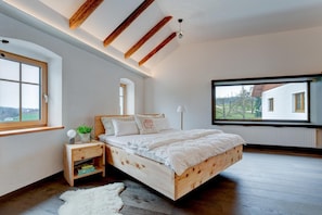 Un dormitorio con cama de madera, una mesita de noche, alfombra blanca, una ventana y vista a una casa.