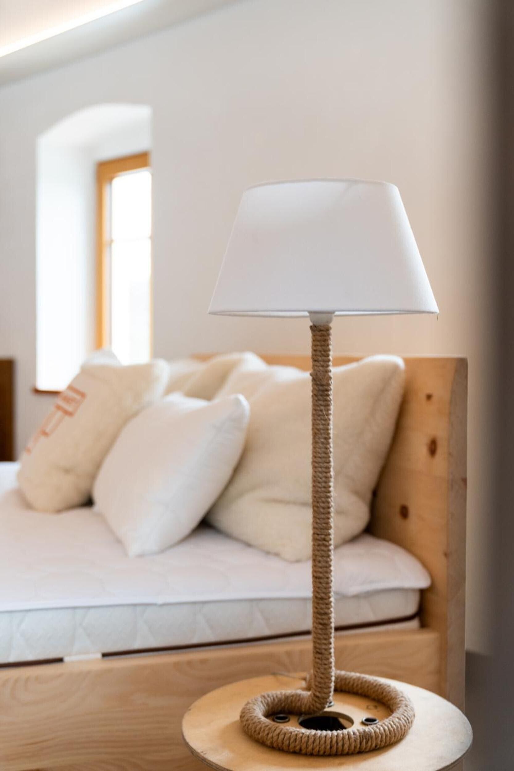 Un árbol para gatos con una lámpara de pantalla blanca, una base redonda de madera y una cama con ropa de cama y almohadas blancas al fondo.