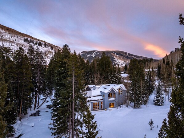 A two-story house surrounded by snow-covered ground and tall pine trees, with a mountain in the background. - 6 Bedroom Riverside Haven in East Vail (Vail)