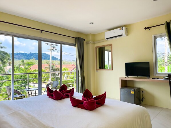 A hotel room with a large bed, a television, and a view of greenery and buildings outside the window.