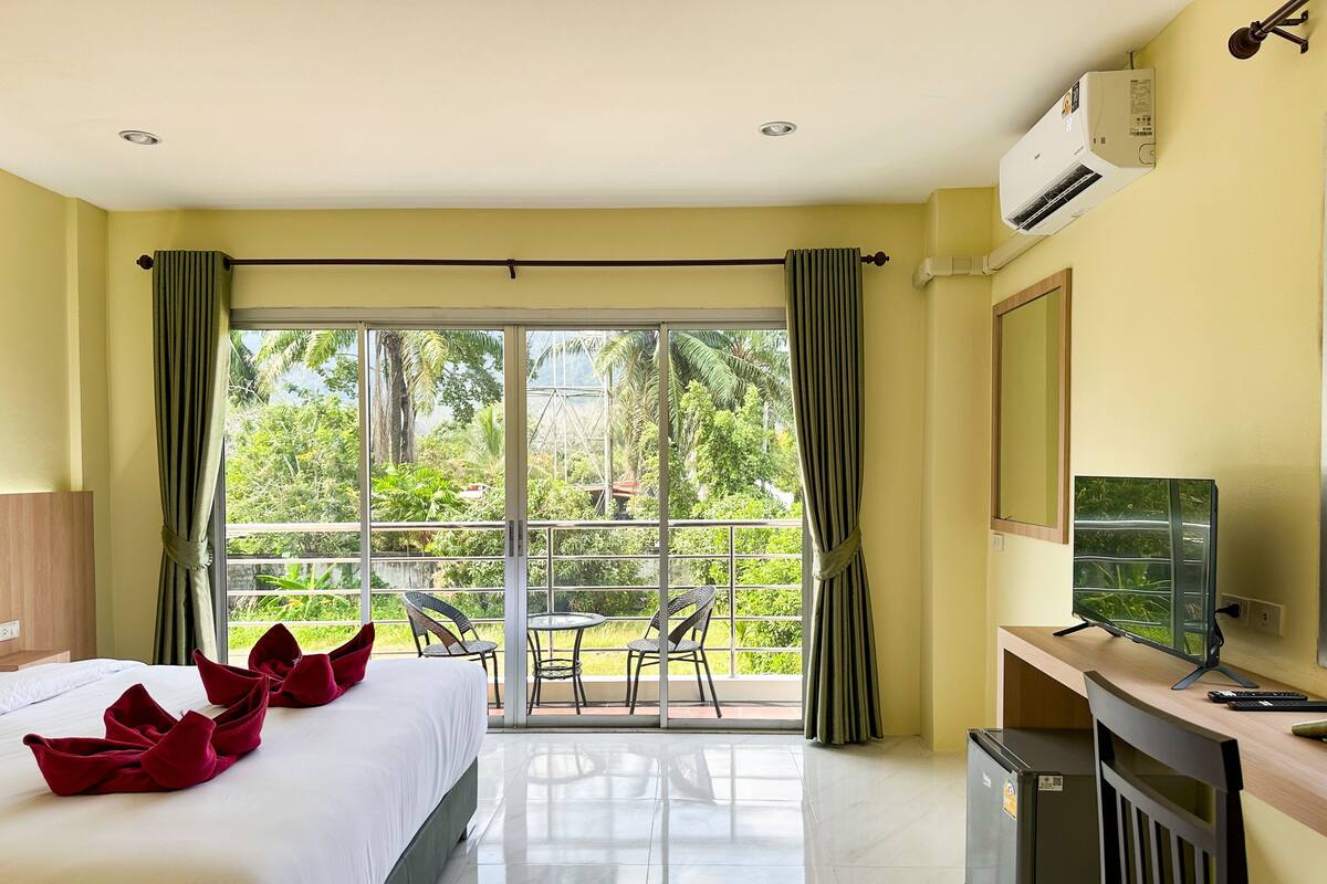 A hotel room with a bed, a desk, a television, and a balcony with a view of greenery.