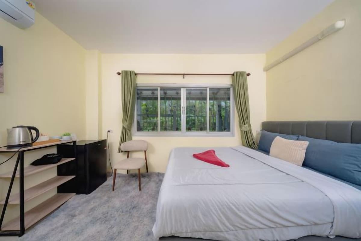 A bedroom with a bed, a desk, a chair, a window with curtains, and an air conditioning unit.