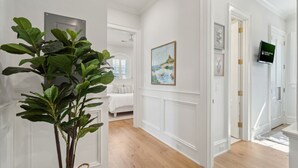 A hallway with white walls, a large potted plant, a painting, and a television on the wall.