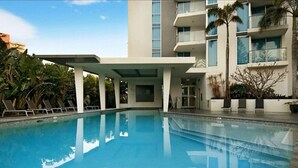A swimming pool with lounge chairs and a covered area, surrounded by a modern building with balconies and palm trees.