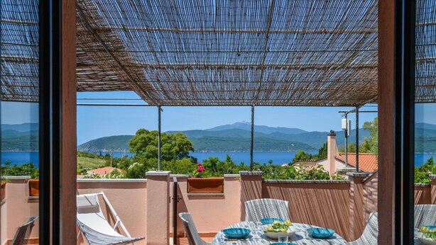 A terrace with a table set for dining, overlooking a scenic view of the sea and mountains.
