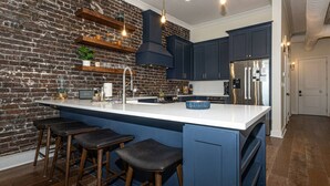 A kitchen with a brick wall, dark blue cabinets, a white countertop, and three bar stools.