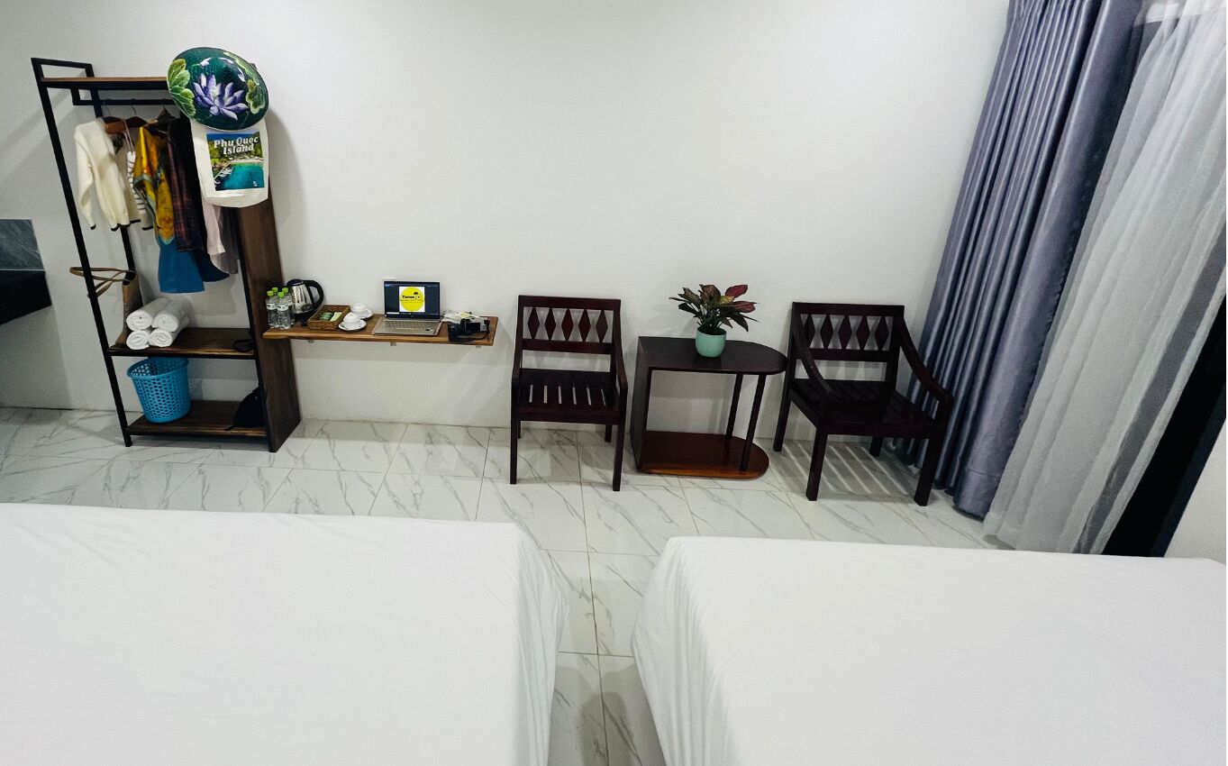 A hotel room with a bed, a desk with a laptop, two chairs, a small table with a plant, and a shelf with towels and a hat.