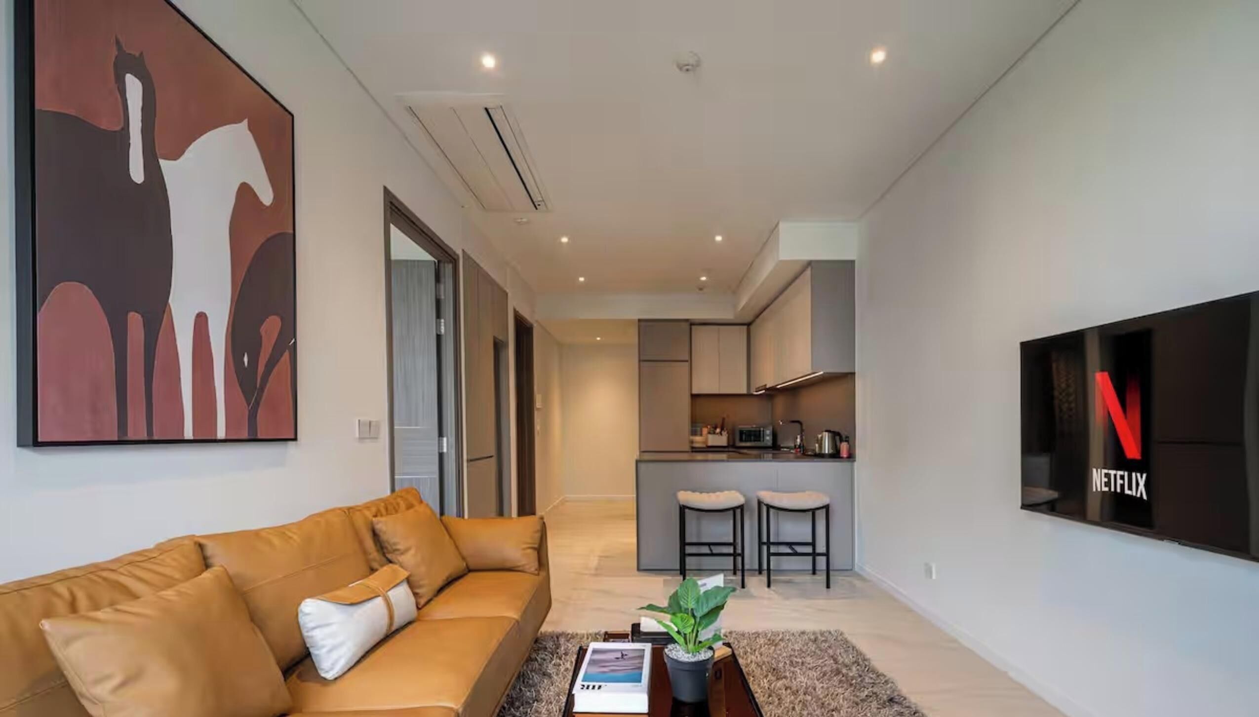 Una sala de estar moderna con un sofá, una mesita de centro con un libro y una planta, una cocina al fondo y un televisor Netflix montado en la pared.