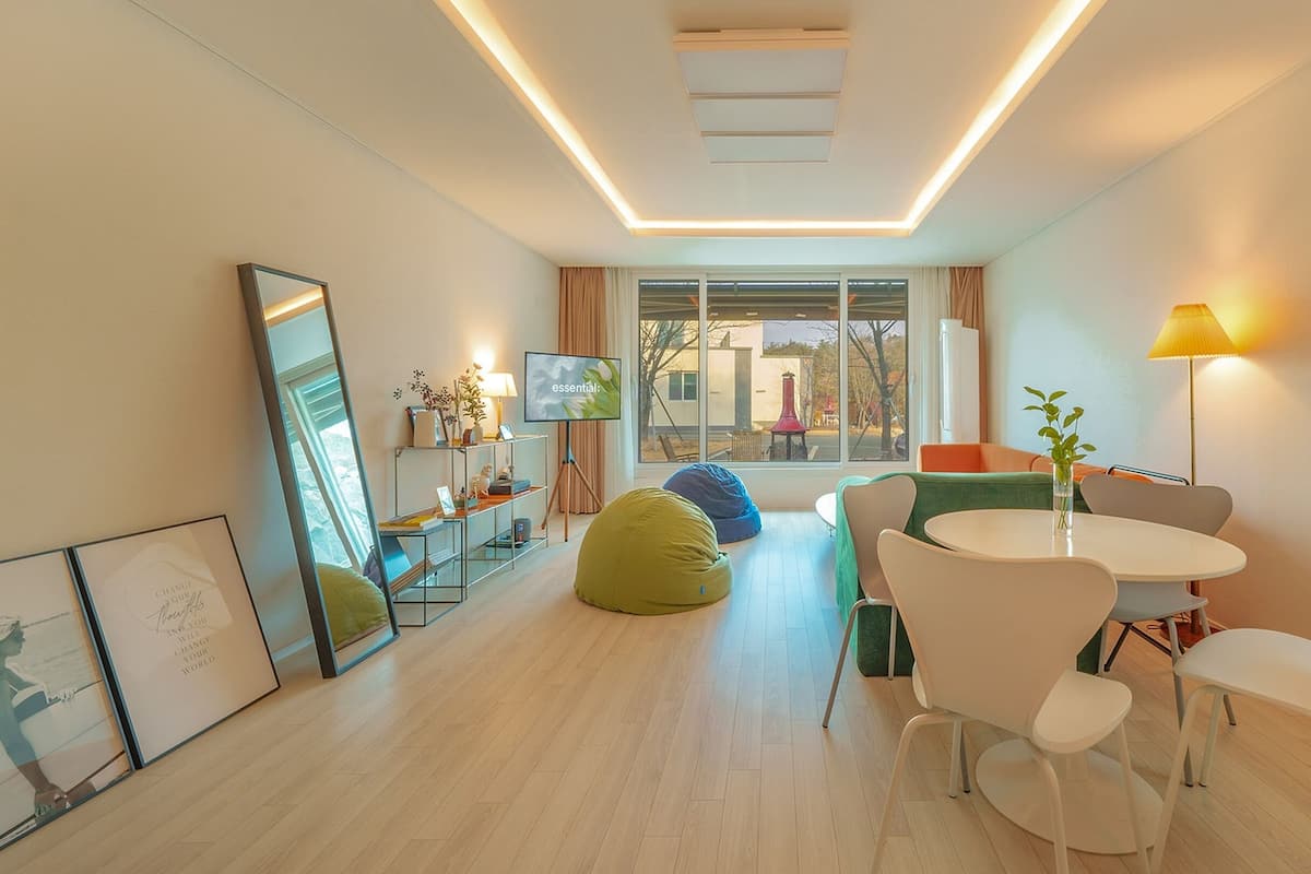 A modern living room with a flat-screen TV, a dining table with chairs, bean bags, and a large window with a view of the outside.