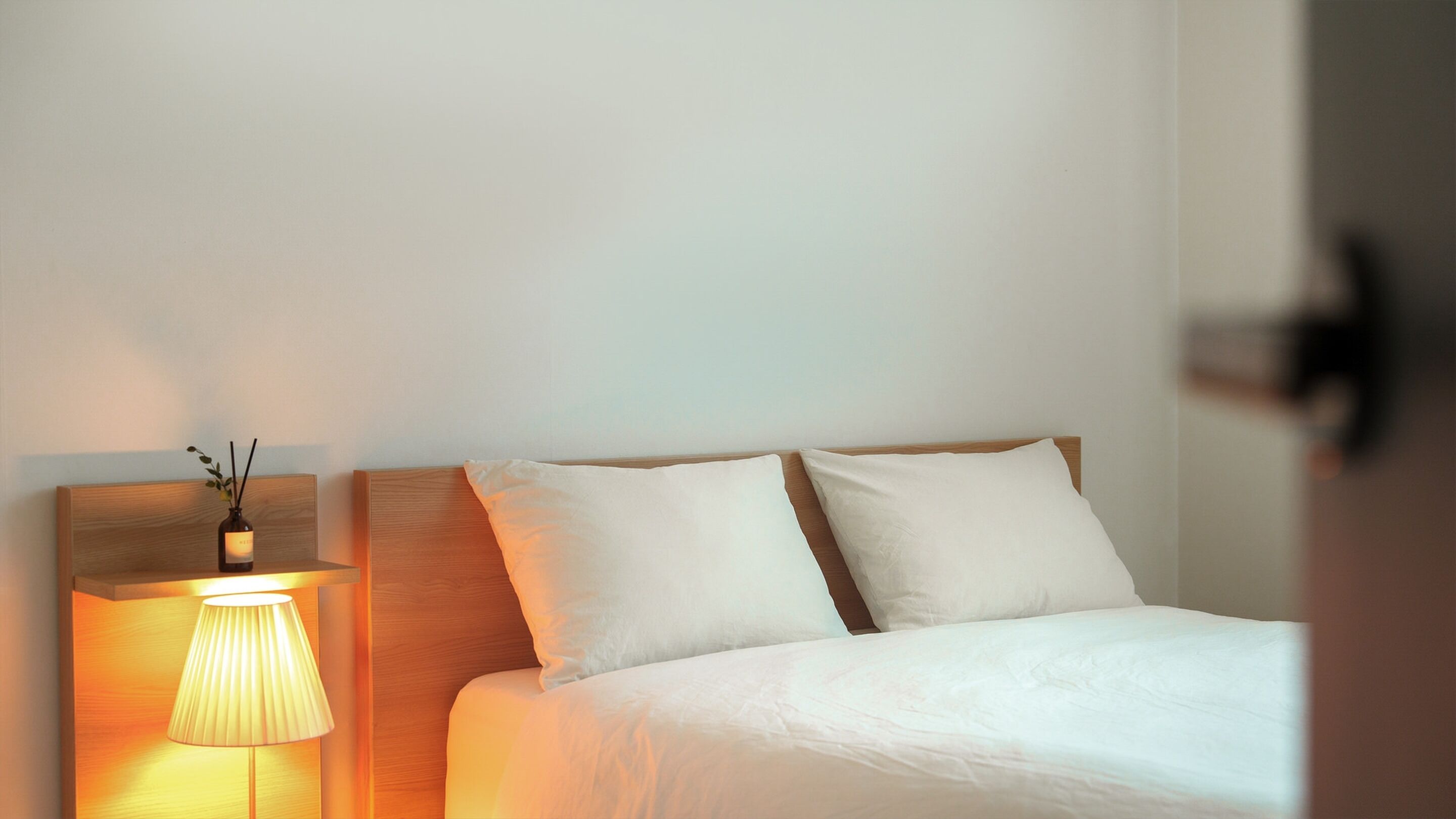 A hotel room with a bed, two pillows, a nightstand with a lamp, and a wall-mounted vase with reeds.