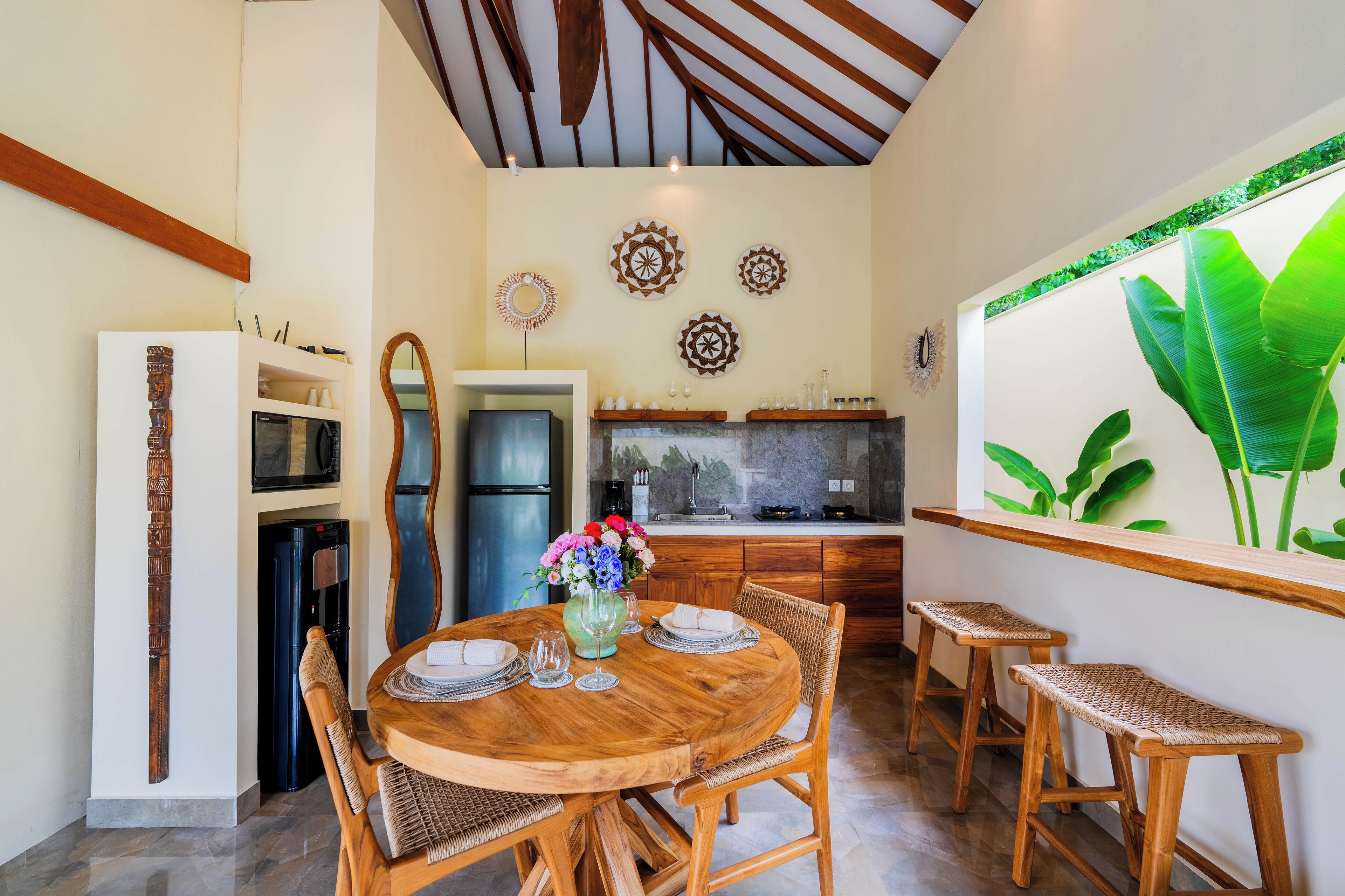 A dining area with a round wooden table, two wicker stools, and a vase with flowers.